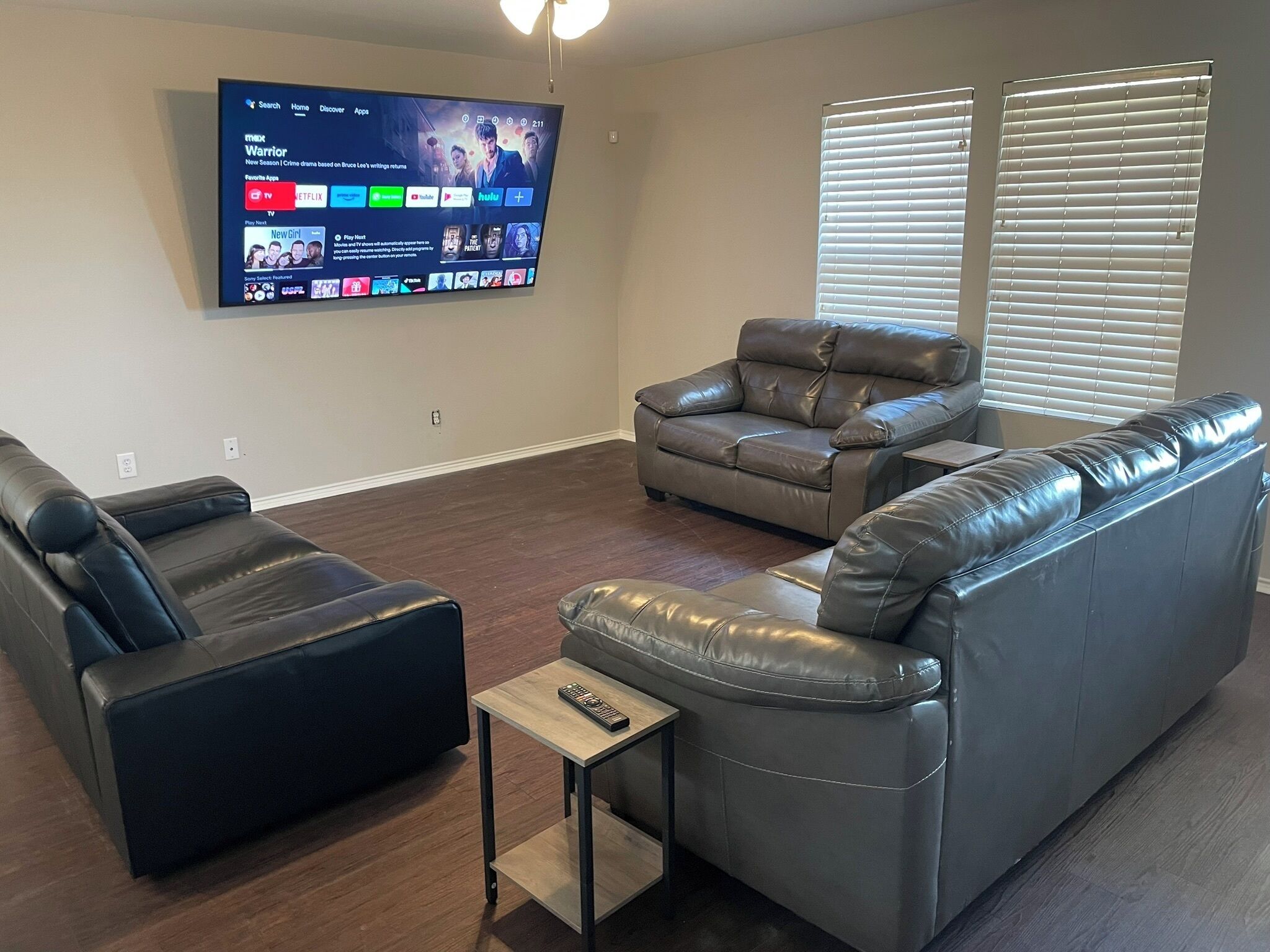 Una sala de estar con un televisor de pantalla plana montado en la pared que muestra la interfaz de un servicio de streaming. Hay dos sofás de cuero y una pequeña mesa de centro de madera con un control remoto encima.