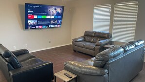 Una sala de estar con un televisor de pantalla plana montado en la pared que muestra la interfaz de un servicio de streaming. Hay dos sofás de cuero y una pequeña mesa de centro de madera con un control remoto encima.