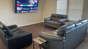 Una sala de estar con un televisor de pantalla plana montado en la pared que muestra la interfaz de un servicio de streaming. Hay dos sofás de cuero y una pequeña mesa de centro de madera con un control remoto encima.