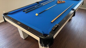 Una mesa de billar con superficie de fieltro azul, tacos, una bola blanca y un juego de bolas de billar.
