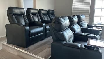 Sistema de cine en casa con cuatro sillones de cuero negro reclinables, piso de madera y ventilador de techo.