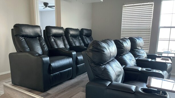 Sistema de cine en casa con cuatro sillones de cuero negro reclinables, piso de madera y ventilador de techo.