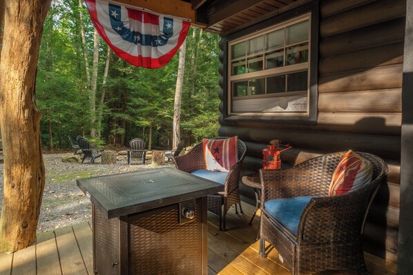 A wooden cabin with a porch, furnished with wicker chairs and a table, and an American flag hanging above.