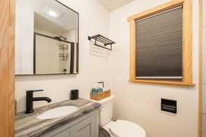 Un baño con lavamanos blanco, un espejo, toallero, una ventana con persianas y un inodoro.