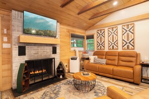 A cozy living room with a fireplace, a large flat-screen TV, a brown leather sofa, and a patterned rug.