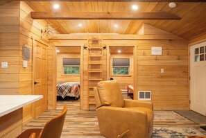 Interior de una cabaña con detalles de madera, una cama, una silla, una mesa y una escalera que lleva a un altillo.