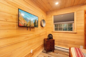 Una habitación con paneles de madera, un televisor montado en la pared, una mesita de noche de madera con un control remoto y una ventana con persianas.