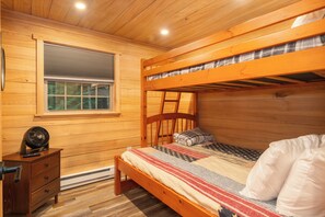Una cama litera con cabecera de madera, una mesita de noche con un ventilador y una ventana con persianas.