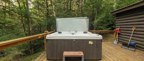Un jacuzzi en una terraza de madera con un banco de madera al frente, rodeado por un bosque.