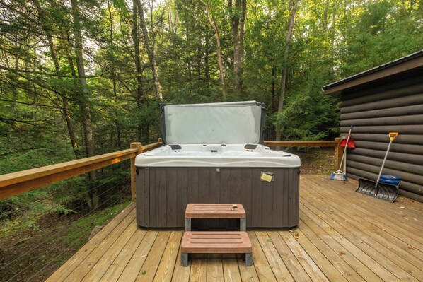 Un jacuzzi en una terraza de madera con un banco de madera al frente, rodeado por un bosque.