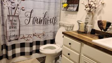 Een badkamer in boerderijstijl, met een zwart-wit geruit douchegordijn, een toilet, een houten wastafelmeubel en een bordje met daarop 'Badkamerregels'.