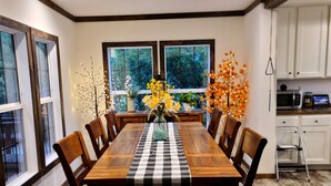 A dining room with a wooden table, chairs, and a checkered tablecloth. There are windows with curtains, a kitchen area with cabinets, and a decorative tree with lights.