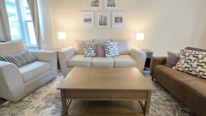 Ein Wohnzimmer mit einem beigen Sofa, einem braunen Sofa und einem Couchtisch aus Holz. Zwei Stehlampen und eine Wand mit gerahmten Bildern sind ebenfalls vorhanden.
