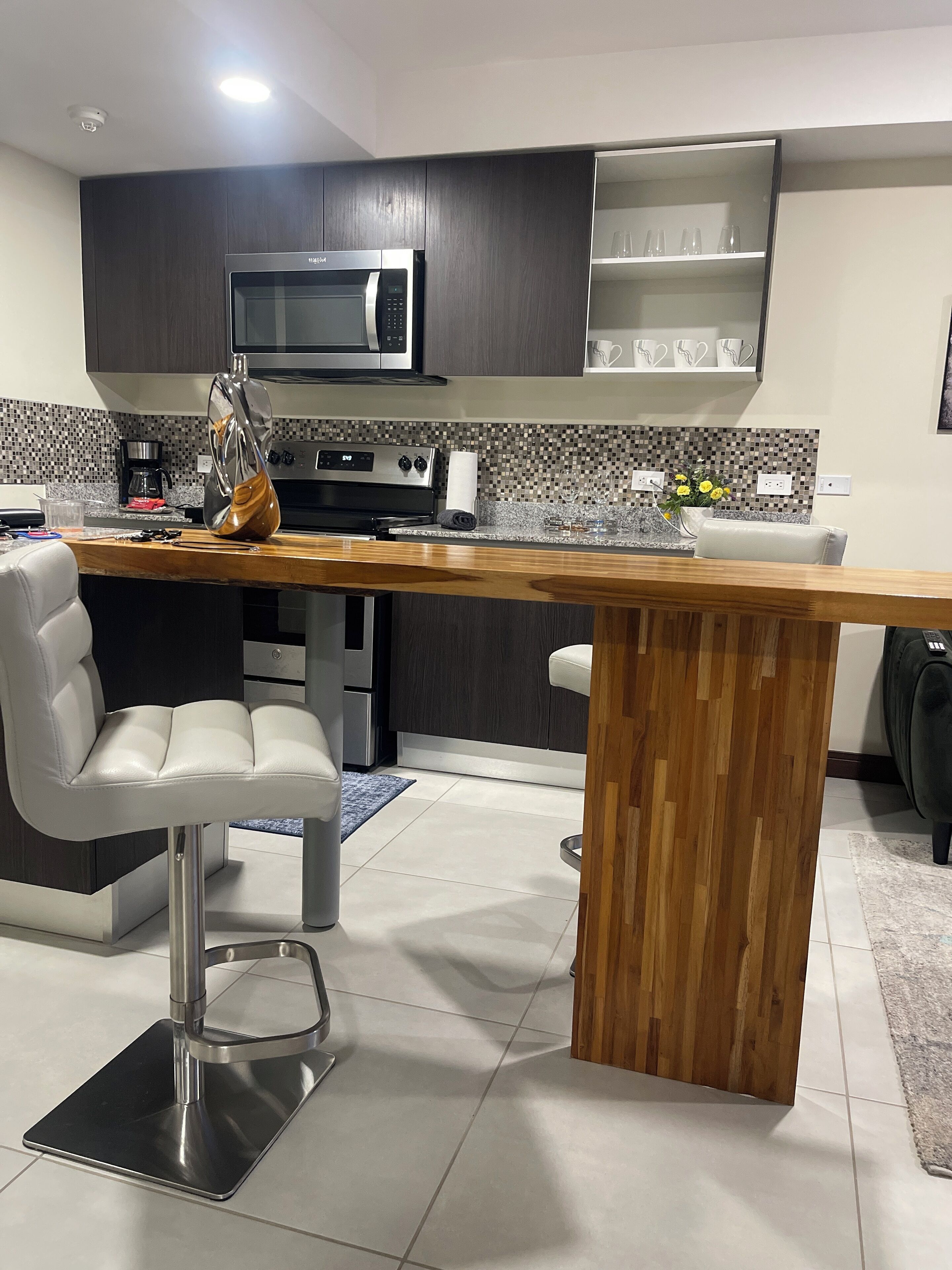 A modern kitchen with dark cabinets, a wooden island, stainless steel appliances, and a seating area with white chairs.