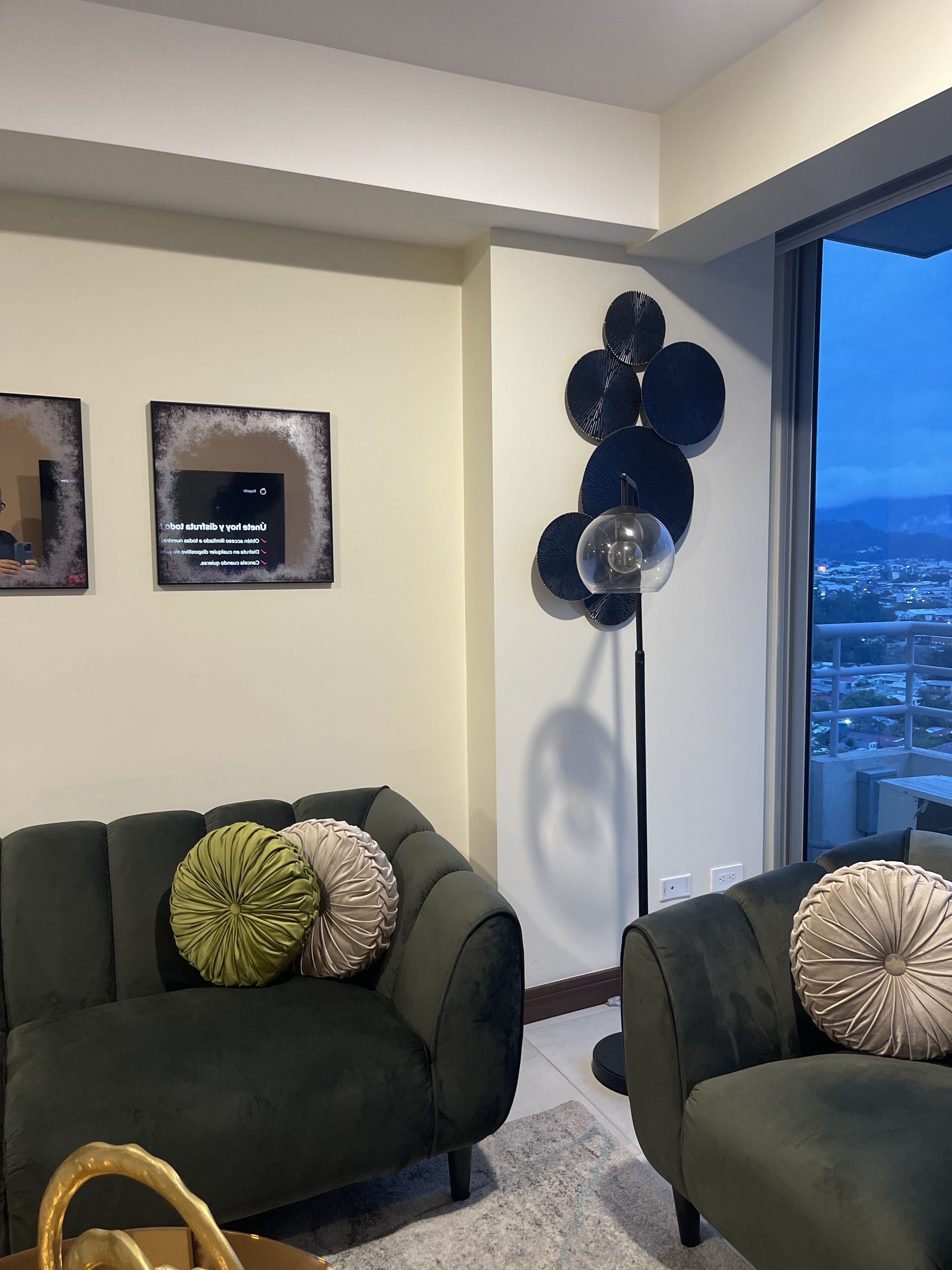 Un salon avec deux canapés vert foncé, un lampadaire et une vue sur la ville, visible à travers une grande fenêtre.