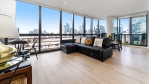A spacious living area with a black leather sofa, a dining table, and chairs. Large windows offer a view of the city skyline. - Docklands Melbourne CBD Views (Docklands)