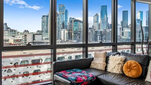 A modern room with large windows, a black leather sofa, and a view of a city skyline. - Docklands Melbourne CBD Views (Docklands)