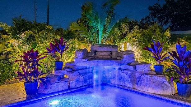 A nighttime outdoor pool area with a waterfall feature, surrounded by potted plants and palm trees. - Stunning 3 BR Oasis with Waterfall Grotto by white sands of Clearwater Beach (Clearwater)