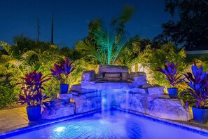A nighttime outdoor pool area with a waterfall feature, surrounded by potted plants and palm trees.
