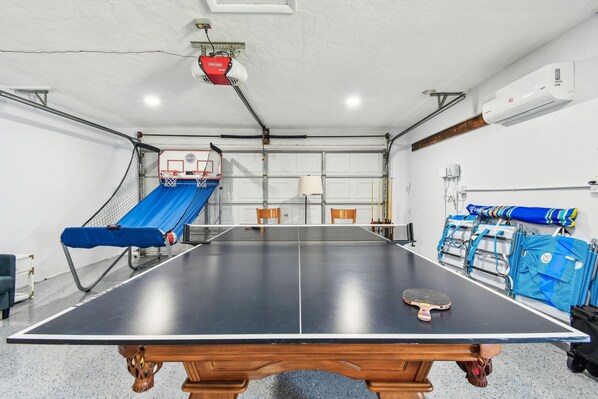 A ping pong table in a garage with a basketball hoop, chairs, and a lamp.
