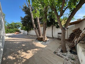 A courtyard with a paved walkway, trees, a white wall, and a stack of firewood.