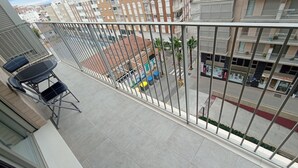Ein Balkon mit Tisch und Stühlen, Blick auf eine Straße mit Gebäuden und Palmen, sowie ein rot und weiß gestrichener Bordstein.