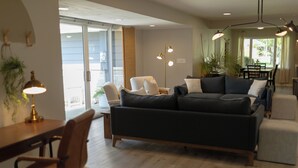 Una sala de estar moderna con un sofá gris oscuro, una mesa de centro de madera y una alfombra blanca. Hay un escritorio con una lámpara y una silla, y un área de comedor con una mesa y sillas.