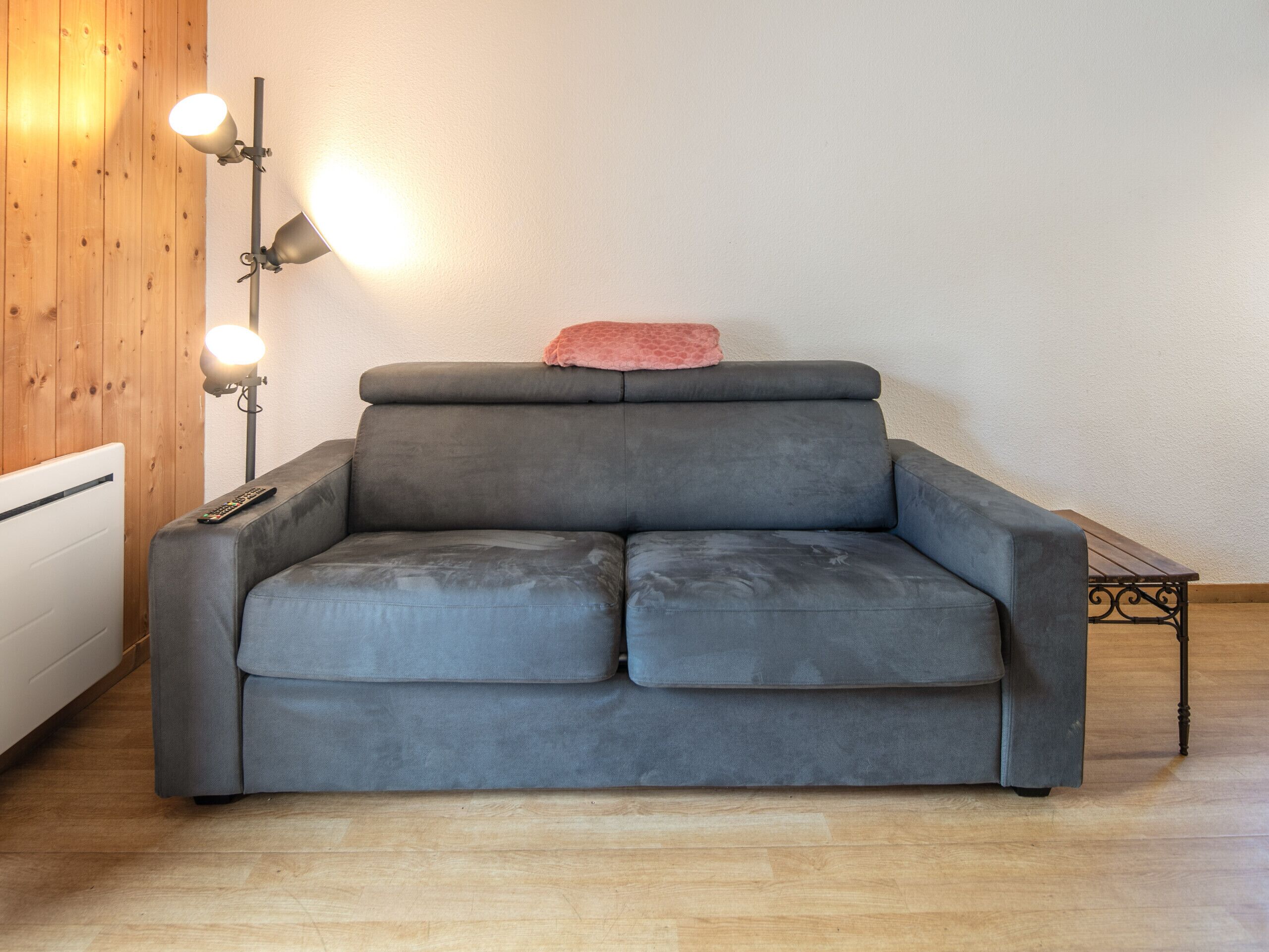 Een grijze bank met een roze kussen, een staande lamp en een radiator in een kamer met houten wanden en een houten vloer.