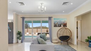 Ein modernes Wohnzimmer mit einem grauen Sofa, einem runden Korbstuhl und Blick auf einen Pool durch große Glasfenstertüren.