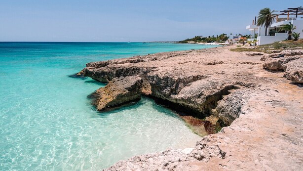 A coastal area with clear turquoise waters, rocky formations, and a modern building with a white facade and palm trees.