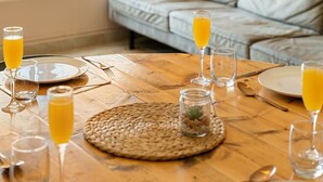 A cozy living room with a sofa, a dining table set with glasses of orange juice, and a large window with a view of greenery.