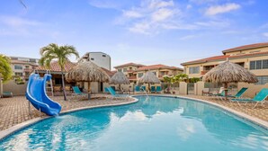 A swimming pool with a blue slide, lounge chairs, and thatched umbrellas.