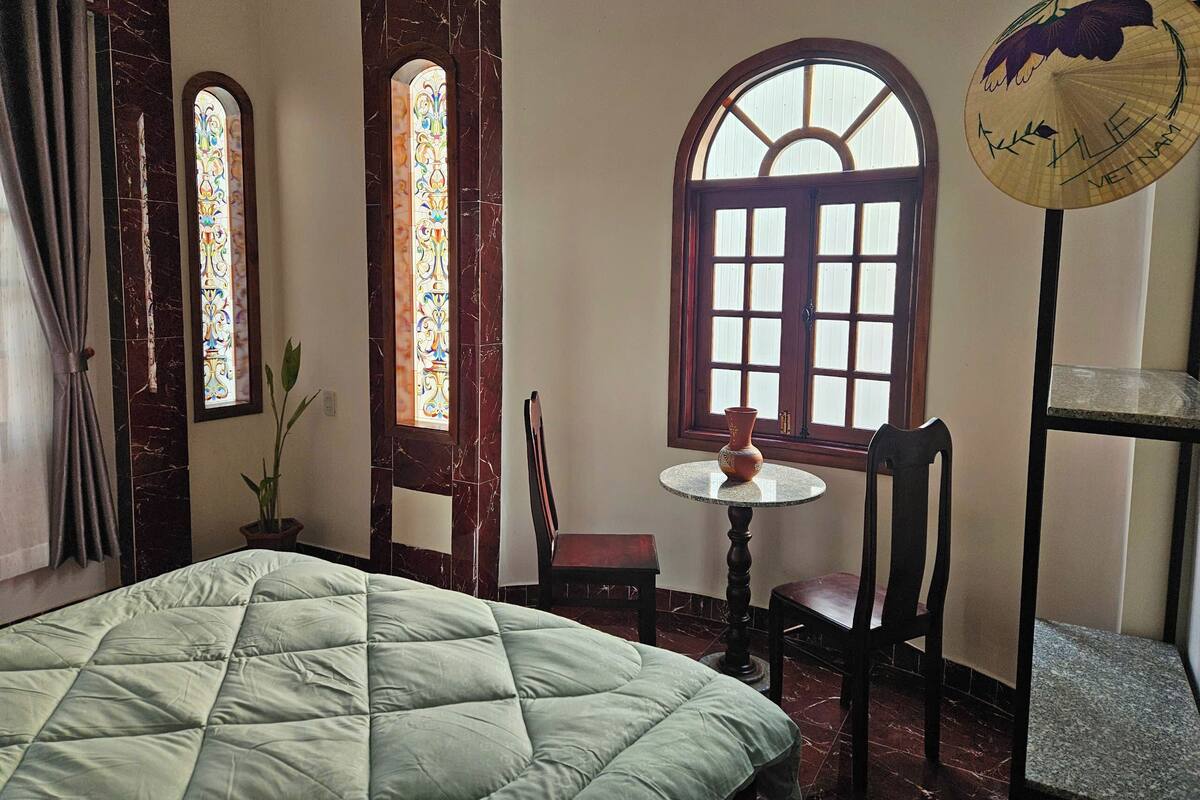 Una camera con un letto, un tavolino con un vaso, due sedie e una finestra con cornice decorativa.