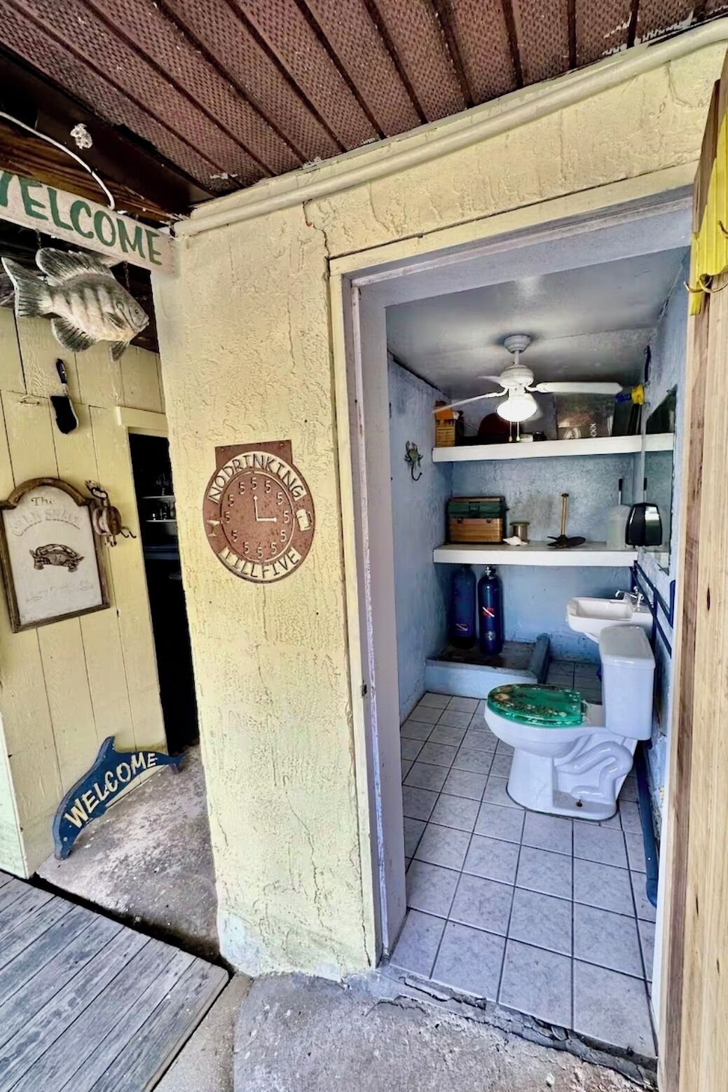 Een kleine, rustieke badkamer met één toilet, een plank met diverse spullen, en een klok aan de muur.