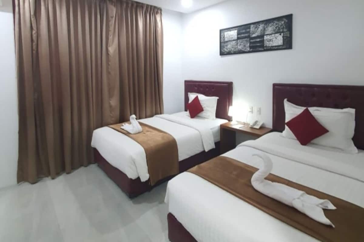A hotel room with two beds, each with a brown headboard, white bedding, and a red pillow. There is a swan-shaped towel on each bed. A nightstand with a phone and lamp is between the beds.