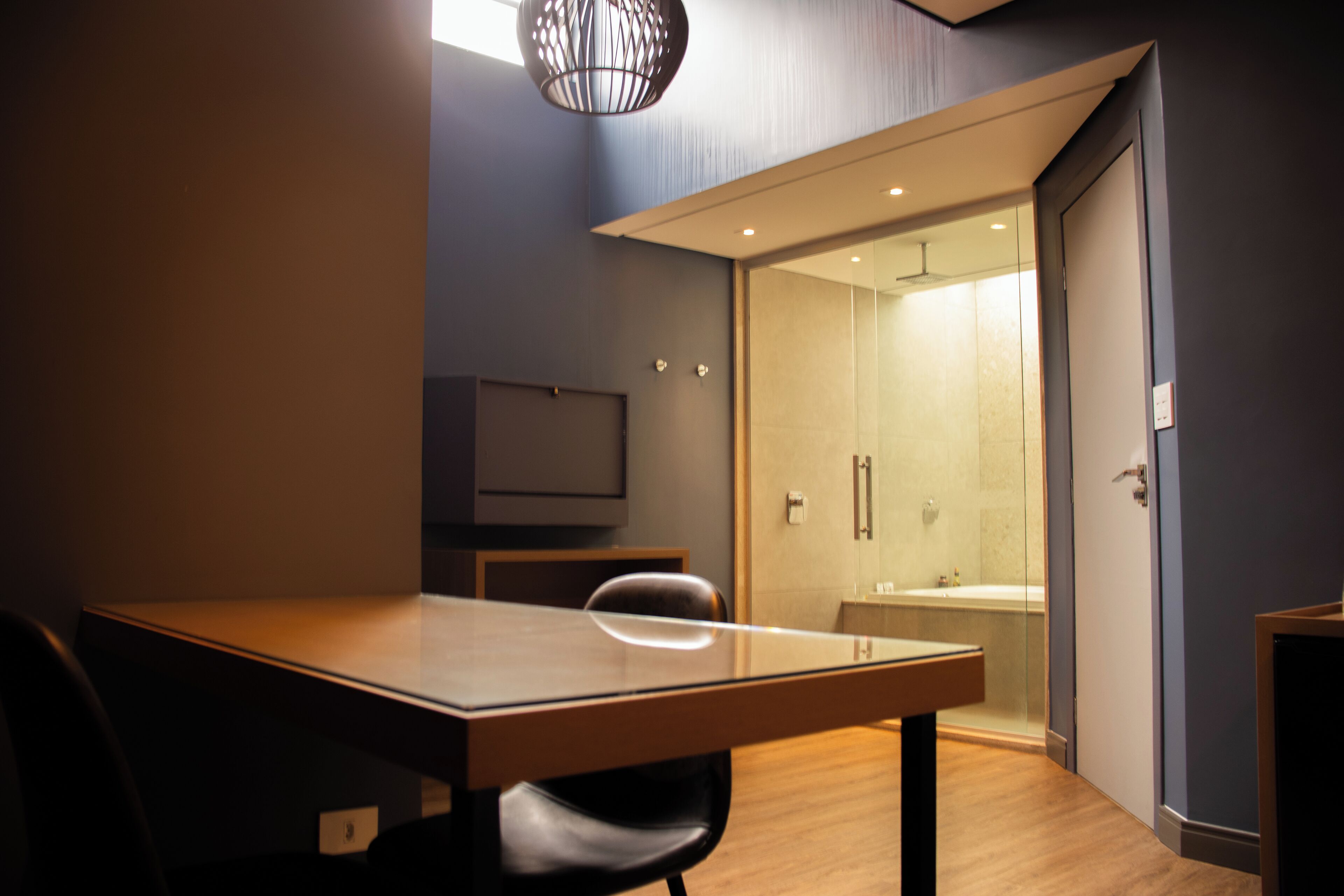 A modern hotel room with a wooden table, a black chair, a glass shower enclosure, and a visible bathroom with a bathtub.
