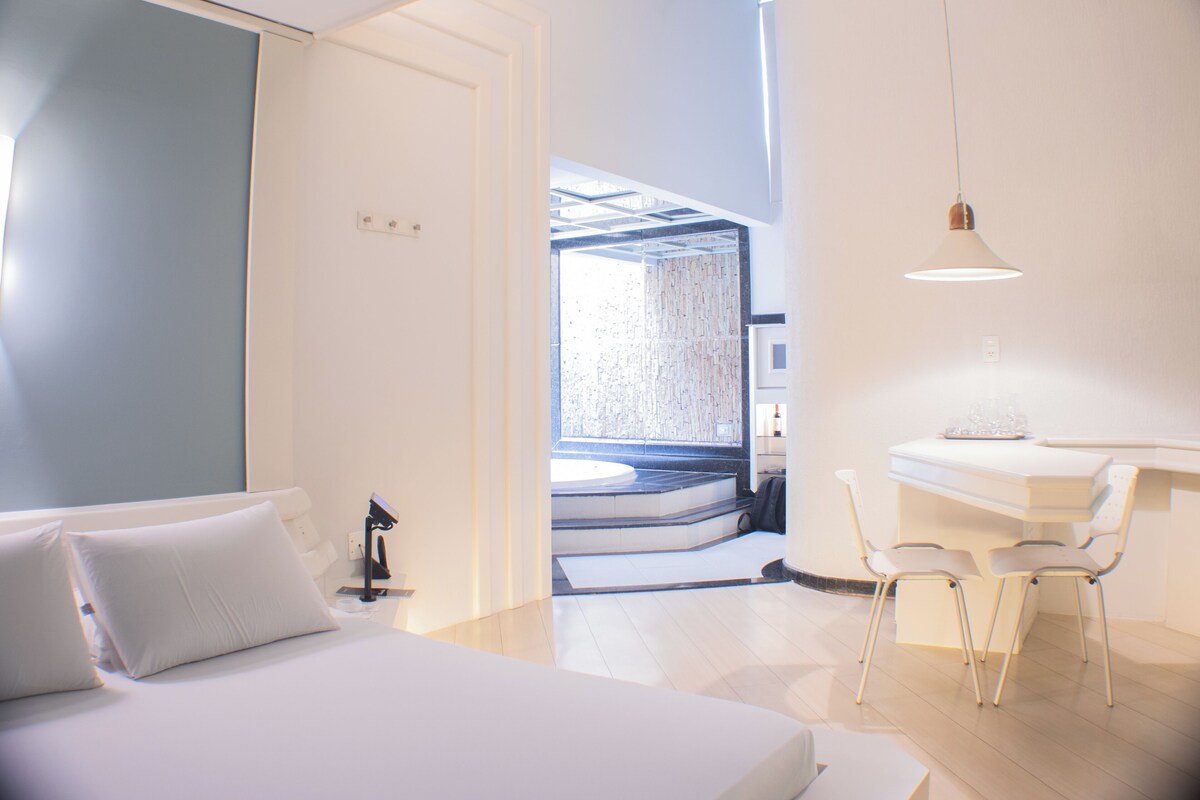 A modern hotel room with a bed, a desk, and a chair. There is a window with a view of a staircase and a wall with a geometric pattern.