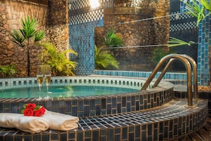 Un jacuzzi con borde de azulejos, rodeado de plantas y un muro de piedra.