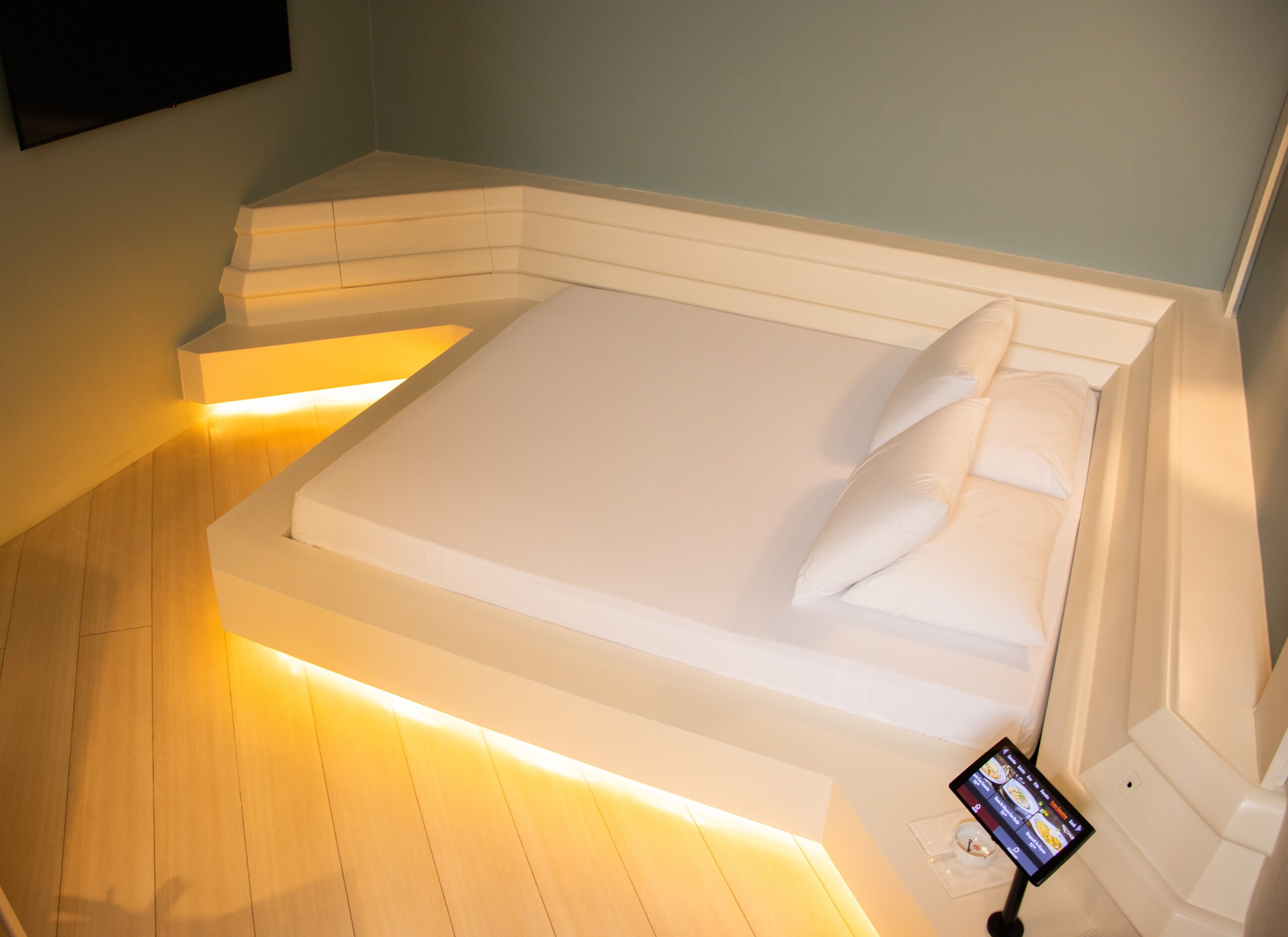 A modern hotel room with a large bed, a bedside table, and a digital device on the nightstand.