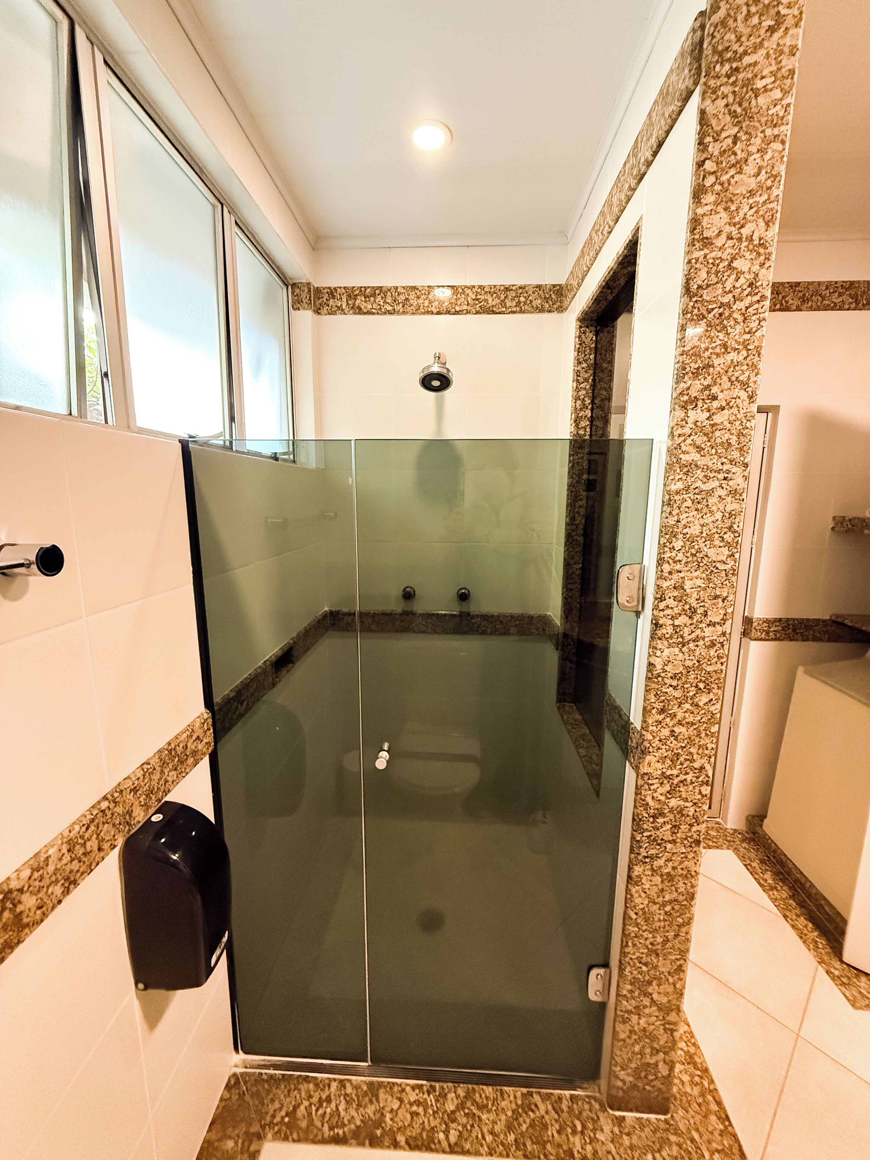 Une douche fermée par des parois de verre, avec un bord en marbre, une pomme de douche visible et un distributeur de gel hydroalcoolique.