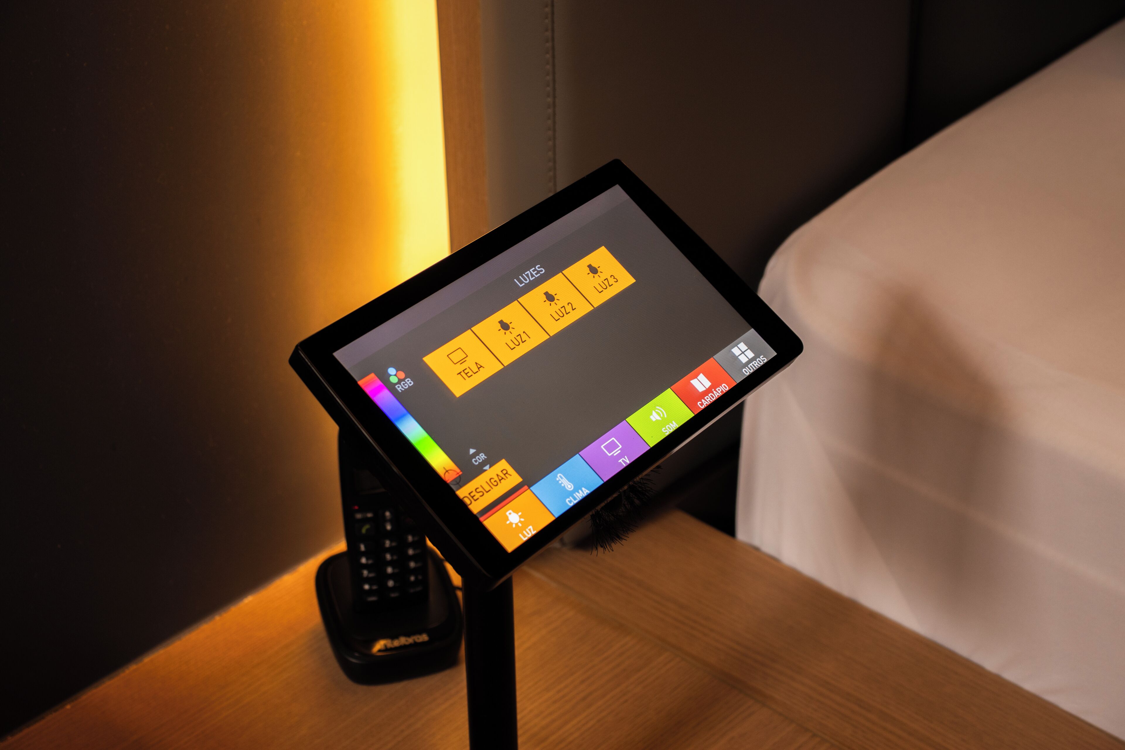 Una tableta muestra una interfaz de control con varios iconos, probablemente para la configuración de la habitación o servicios del hotel, colocada sobre un soporte junto a un teléfono.