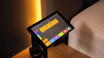 Una tableta muestra una interfaz de control con varios iconos, probablemente para la configuración de la habitación o servicios del hotel, colocada sobre un soporte junto a un teléfono.