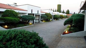 Um caminho conduz a uma fileira de edifícios modernos, com sebes bem aparadas e pequenas luzes ao longo do percurso.
