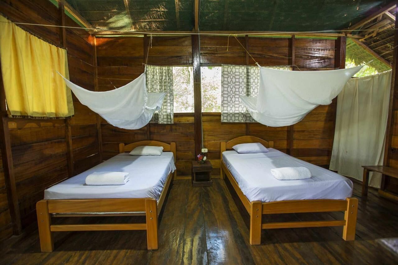 Una habitación con dos camas de madera, una mesita y un ventanal con cortinas.