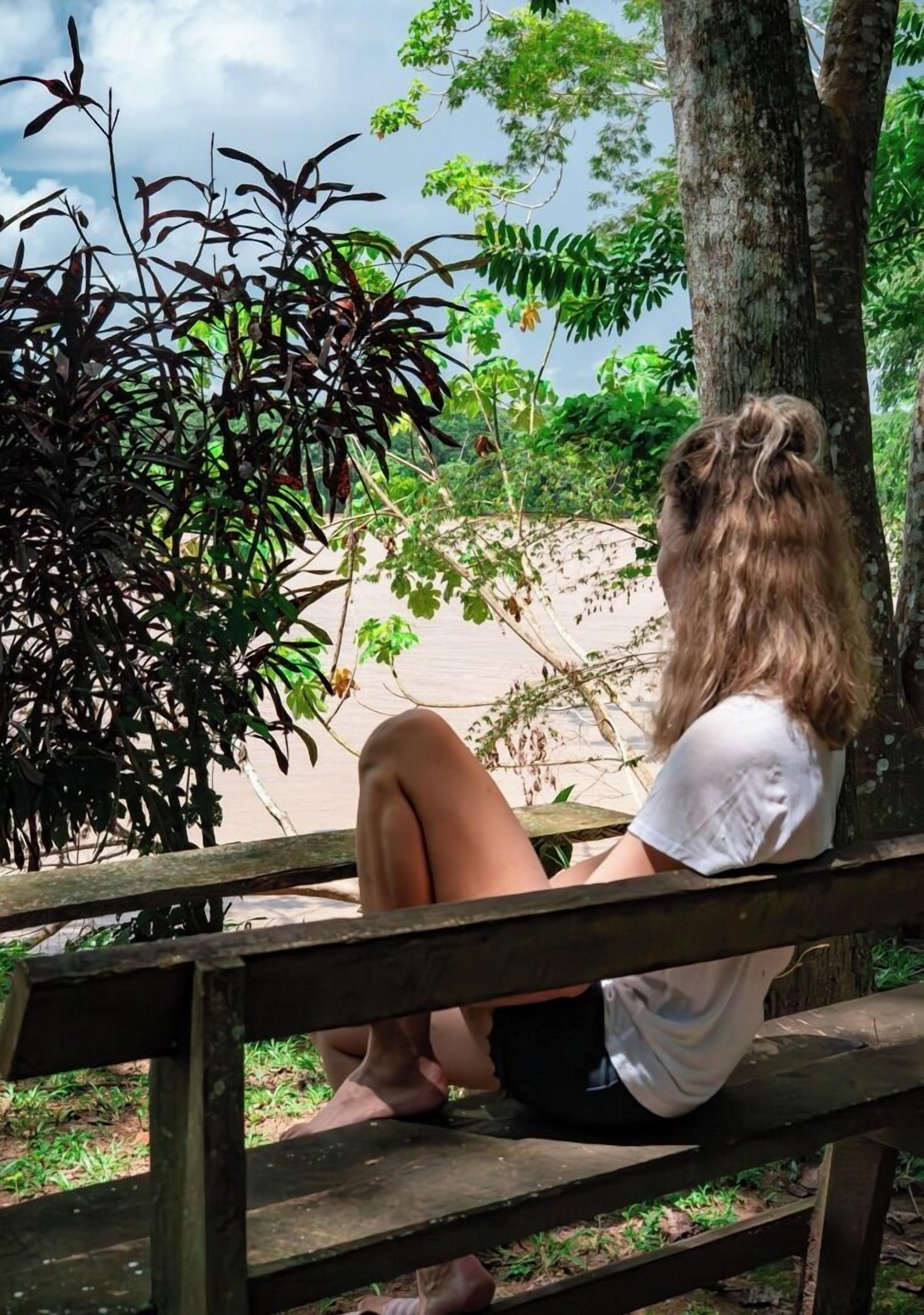 A person sitting on a wooden bench outdoors, surrounded by greenery and trees.