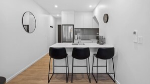 A modern kitchen with white cabinets, a stainless steel refrigerator, and a coffee maker. There are three black bar stools at the counter. - Ride Recover & Rest | Arthurs Point Stay Near Ski & Hot Pools (Queenstown)
