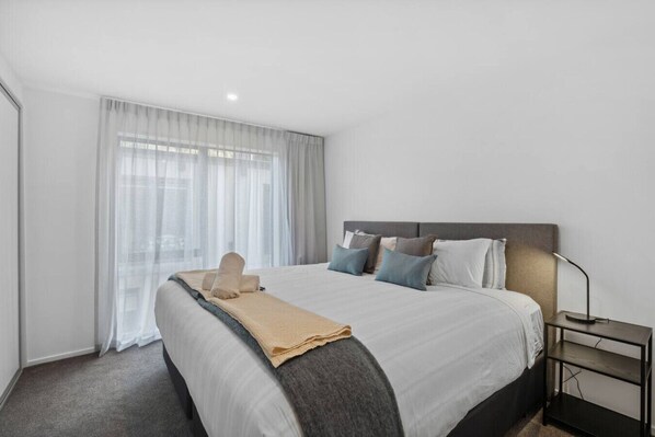 A modern hotel room with a large bed, a bedside table with a lamp, and sheer curtains on the window. - Ride Recover & Rest | Arthurs Point Stay Near Ski & Hot Pools (Queenstown)