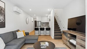 A modern living room with a grey sofa, a coffee table, and a TV on a wooden stand. There is a kitchen area with a fridge and a dining table with chairs. A staircase leads to another level. - Ride Recover & Rest | Arthurs Point Stay Near Ski & Hot Pools (Queenstown)