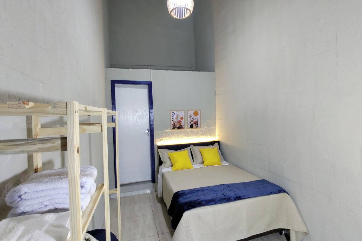 A small, compact room with a bunk bed, a single bed with yellow pillows, a bedside table with a lamp, and a door with a blue frame.