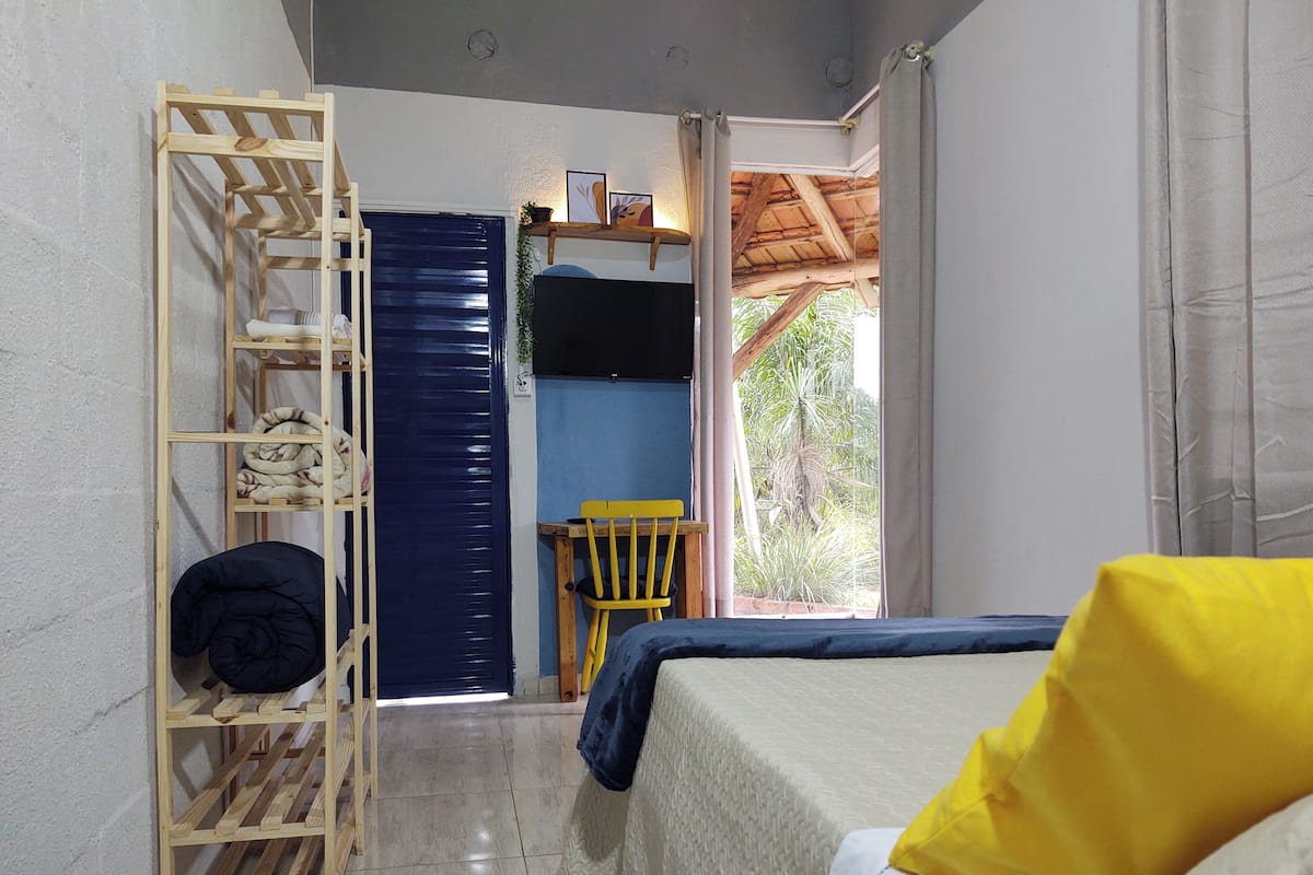 A bedroom with a bed, a wooden shelf unit, a chair, and a view of greenery outside.